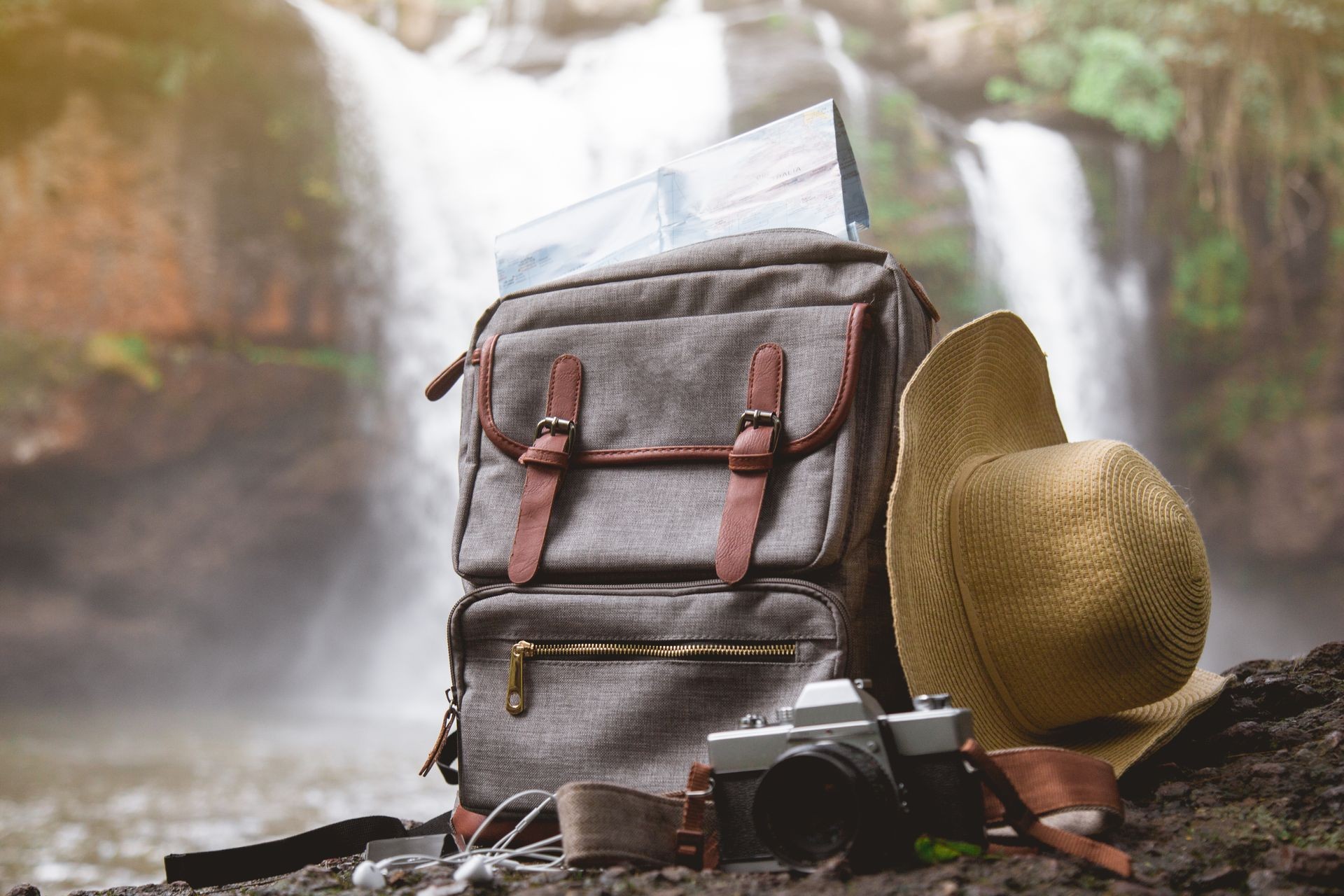 Map in Backpack vintage, Mobile phone with earphone, film camera and hat at amazing background waterfall and forest with a traveler at sunset. Travel and hipster concept.